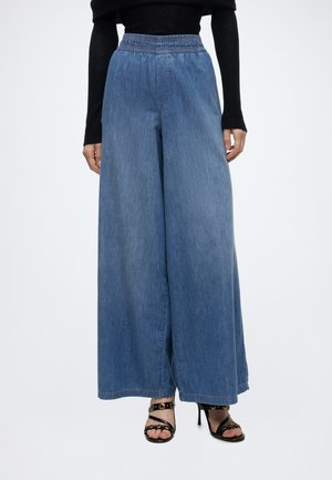 Kobieta nosząca niebieskie, szerokie spodnie denim z wysokim stanem, w połączeniu z czarną bluzką z długim rękawem oraz czarnymi sandałami na wysokim obcasie z paskami wokół kostki.
