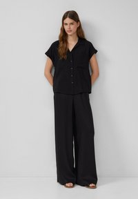 Schwarzes, übergroßes Button-Up-Hemd mit zwei Brusttaschen, kombiniert mit weiten schwarzen Hosen, die beide aus leichtem Stoff gefertigt sind.