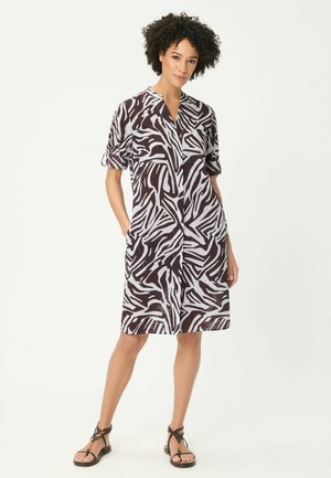 Frau mit lockigem Haar, die ein knielanges schwarz-weißes Zebra-Muster-Kleid und schwarze Riemchensandalen trägt, steht mit den Händen in den Taschen.