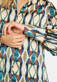 Camisa de patrón multicolor con diseño geométrico que presenta azules, verdes y marrones, con botones de perla y mangas fruncidas en la muñeca.