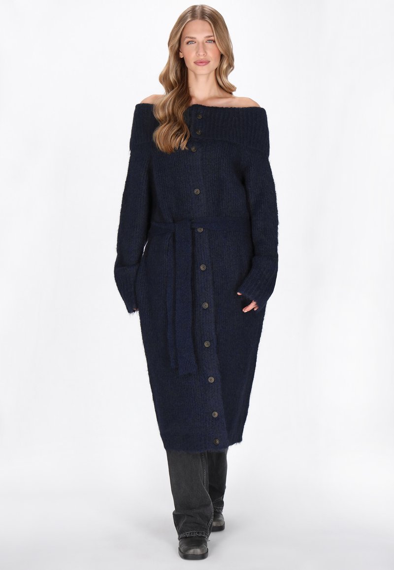 Tengerészkék, vállnélküli kötött ruha hosszú ujjal, gombos elülső résszel és megkötős övvel. Texturált szövet és bordázott nyak, teljes hosszúságú sziluett.