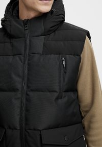 Gilet imbottito nero con colletto alto, chiusura lampo frontale e una piccola tasca sul petto. Presenta un tessuto testurizzato e un contrasto di beige sottostante.