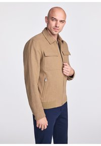 Jasnobrązowa kurtka z zapięciem na zamek, dwiema kieszeniami na piersi, bocznymi kieszeniami na zamek i ściągaczem na dole. Gładka faktura materiału i wyraźna struktura.