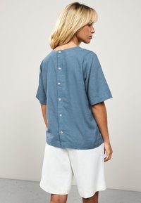 Light blue, short-sleeve shirt with a subtle texture and a round neckline, featuring a button-down back detail and a relaxed fit.
