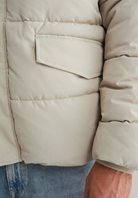 Chaqueta acolchada beige con un bolsillo lateral, textura suave y puños acanalados, combinada con denim claro.