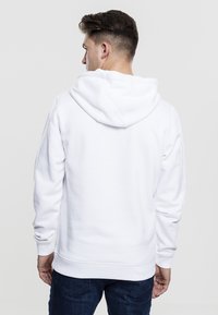 Sudadera blanca con capucha, de corte holgado, puños y dobladillo de canalé, textura suave y una gran capucha ajustable. Se muestra la vista posterior.