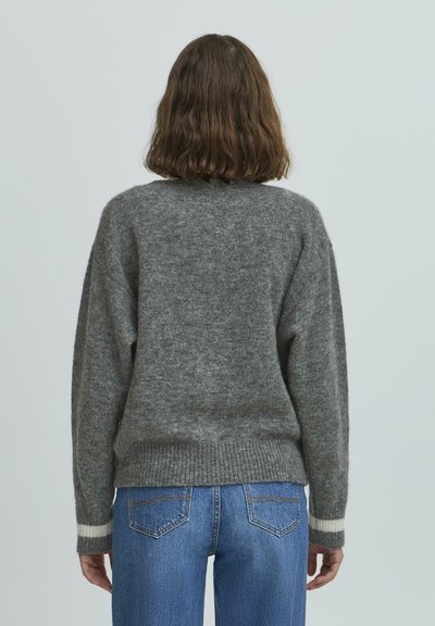 Sivý pletený sveter s voľným strihom, rebrovaným lemom a manžetami, so bielymi pruhmi na manžetách. Nosený s modrými džínsami.