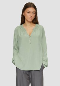 Chemise verte clair à col rond, à manches longues, avec des détails froncés aux épaules. Associée à un pantalon gris.