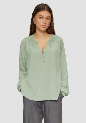 Chemise verte clair à col rond, à manches longues, avec des détails froncés aux épaules. Associée à un pantalon gris.