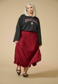 Schwarzes langärmliges Shirt mit "Chicago" Grafik, kombiniert mit einem plissierten bordeauxfarbenen Rock. Schwarze, spitz zulaufende Schuhe runden das Outfit ab.