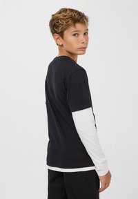 Czarna koszulka z krótkim rękawem nałożona na długą białą koszulę, noszona z ciemnymi spodniami. Materiał wydaje się miękki i ma luźny krój.