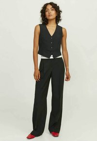 Μαύρο αμάνικο γιλέκο με κοφτή ραφή και κουμπιά μπροστά, συνδυασμένο με φαρδιές μαύρες παντελόνες. Κόκκινα παπούτσια ολοκληρώνουν το σύνολο.