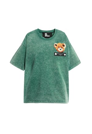 La camiseta verde de gran tamaño presenta un aplique de osito de peluche en el pecho y una etiqueta debajo. Mezcla de algodón con un acabado suave y texturizado.