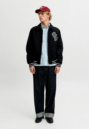 Jeune homme portant une veste universitaire noire, une chemise bleu clair, un jean large foncé avec des revers, des chaussures noires et une casquette de baseball rouge regardant sur le côté.