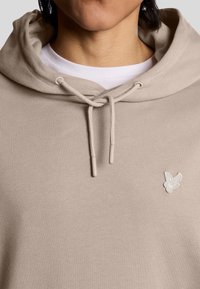 Beige huvtröja med dragsko, med en liten broderad logotyp på bröstet och ett mjukt, texturerat tyg.