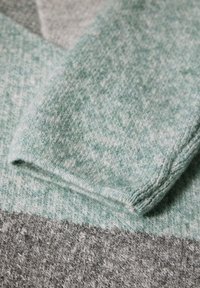 Gebreide trui met een mintgroene en grijze kleurenpalet, textuurstof en een opgerolde mouw die het zachte materiaal benadrukt.