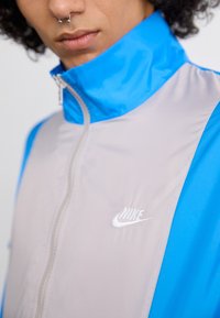 Coupe-vent Nike avec un corps gris clair et des accents bleus, col haut, zip complet et détail de logo blanc sur la poitrine.