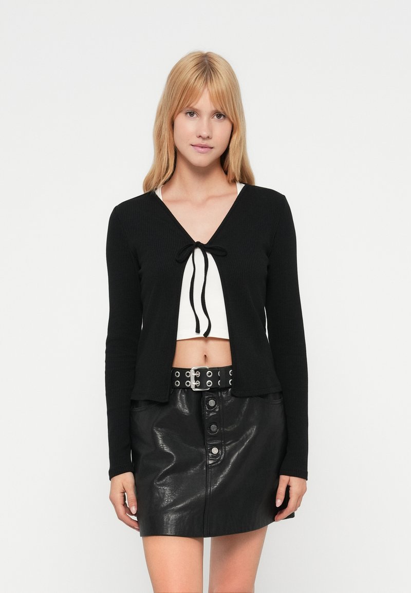 Μαύρο πλεκτό ζακέτα με ριμπ ύφασμα, μακριά μανίκια, V-λαιμόκοψη και δέσιμο μπροστά. Φορεμένο πάνω από λευκό κροπ τοπ, συνδυασμένο με μαύρη δερμάτινη φούστα με κουμπιά μπροστά.