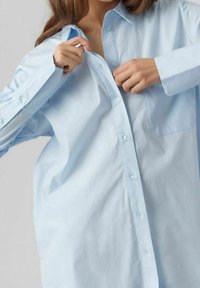 Camicia azzurro chiaro, oversize, con bottoni, realizzata in tessuto morbido, caratterizzata da una singola tasca sul petto e maniche lunghe con polsini abbottonati.