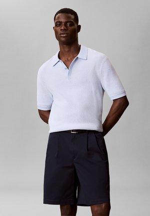 Mężczyzna noszący jasnoniebieską dzianinową koszulkę polo z krótkim rękawem i granatowe plisowane spodenki, stojący z rękami za plecami.