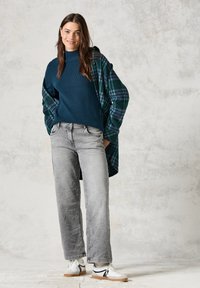 Mörkt teal ribbat tröja i lager med en grön och blå rutig skjorta, kombinerat med ljusgrå vida jeans och vita sportskor.