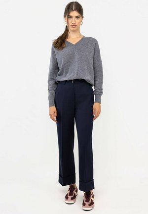 Mujer con el pelo castaño recogido en una coleta, con un suéter gris de cuello en V, pantalones anchos azul marino con puños y zapatillas rosas y marrones, de pie.