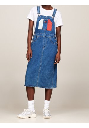 Osoba ubrana w niebieską sukienkę ogrodniczkową z dżinsu z naszywką w kolorach czerwonym, białym i niebieskim na przodzie, białą koszulkę, białe skarpetki i białe tenisówki, stojąca na jednolitym tle.