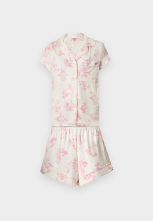 Jeune femme debout pieds nus portant un ensemble de pyjama en satin blanc à manches courtes avec imprimé floral rose sur fond uni.