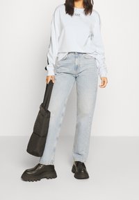 Donna che indossa una maglietta bianca a maniche lunghe EA7, jeans azzurri chiari, scarpe chunky nere, mentre tiene una borsa a tracolla imbottita nera su uno sfondo bianco semplice.