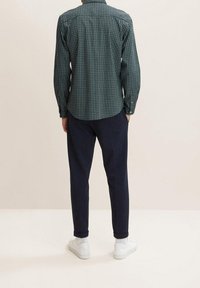 Camicia a quadri verdi con colletto e polsini a bottone; abbinata a pantaloni blu navy e sneakers bianche. L'outfit presenta una vestibilità rilassata e una texture morbida.
