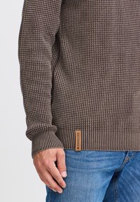 Brązowy sweter dzianinowy z fakturowanym wzorem, ściągaczami przy mankietach i u dołu. Skórzana naszywka oznaczona "JEANSCODE" na dolnym lewym rogu. Noszony z niebieskimi dżinsami.