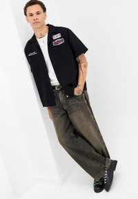Sort kortærmet skjorte med lapper, båret over en hvid t-shirt. Stor, falmet brun jeans og sorte snørebåndsstøvler med sølvdetaljer.