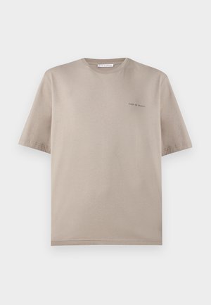 Mladý muž s kučeravými vlasmi má na sebe taupe tričko s krátkym rukávom, biele široké nohavice a hnedé topánky, stojí s rukami v vreckách.