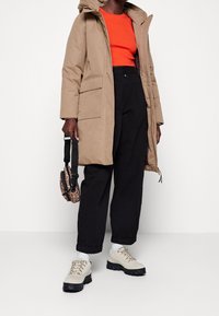 Tan hætteparka med store frontlommer, parret med en lys orange top, sorte bukser og beige snørebåndssko med solide såler.