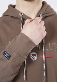 Haut à capuche marron en tissu texturé, équipé d'un cordon de serrage avec un motif noir et blanc, logo brodé et ligne d'accent rouge.