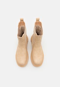 Bottines beiges en faux daim avec une texture lisse, des panneaux latéraux élastiques et des languettes de tirage. Semelles en caoutchouc pour la traction.