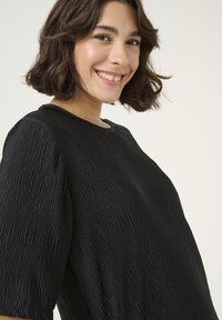 Haut noir texturé avec un col rond et des manches courtes, présentant des motifs à côtes verticales et une coupe ample. Texture de tissu lisse.