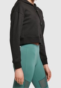 Felpa nera corta con cappuccio regolabile, abbinata a leggings teal a vita alta con diverse trame e dettagli in rete.