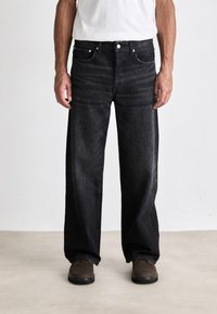 Schwarzweisse Jeans mit weitem Bein und glatter Textur, zwei vorderen Taschen und Knopfverschluss. Kombiniert mit einem schlichten weißen T-Shirt und braunen Schuhen.
