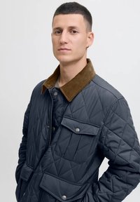 Joven con cabello corto y oscuro, vistiendo una chaqueta de navegante acolchada con cuello de pana marrón, de pie frente a un fondo claro.