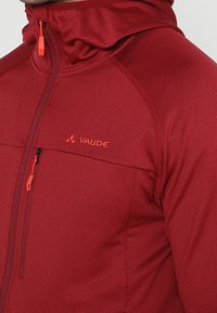 Chaqueta ligera roja con cierre frontal, cuello alto y capucha. Cuenta con un bolsillo en el pecho y cremalleras naranjas en contraste. Logo de la marca visible.