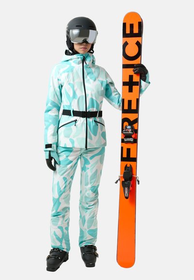 Skiausrüstung in Minzgrün mit weißem Camouflage-Muster, schwarze Handschuhe und Helm; hält einen orangefarbenen Ski mit schwarzer Schrift. Schwarze Skischuhe.