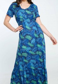 Femme portant une robe bleue à manches courtes avec des motifs de feuilles tropicales vertes et blanches, posant avec une main sur la hanche devant un fond uni.
