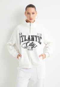 Sudadera blanca con cremallera y estampado gráfico negro que incluye el texto "Atlantic City". Material de algodón suave, corte holgado, puños acanalados y bolsillos laterales.