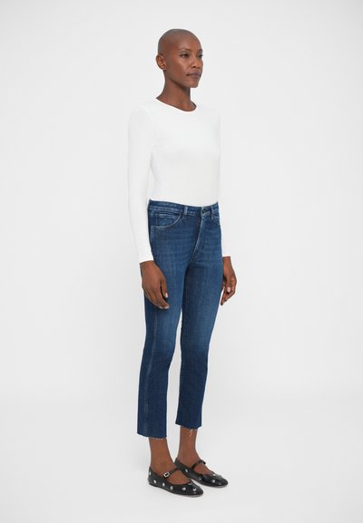 Donna che indossa una maglietta bianca a maniche lunghe, jeans blu a vita alta con orli sfilacciati e scarpe Mary Jane nere con occhielli argentati, in piedi di lato.