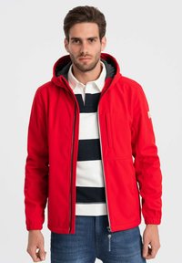 Rød softshell-jakke med hætte, lynlås foran og en brystlomme. Kontrastfor indvendigt og elastiske manchetter, parret med en marineblå og hvid stribet skjorte under.