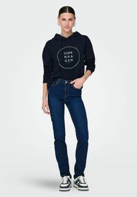 Felpa blu marina con grafica "Copenaghen", abbinata a jeans blu scuro. Il modello indossa sneakers con strisce bianche e nere, su uno sfondo neutro.
