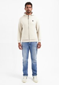 Crème hoodie met textuurstof en rits aan de voorkant, gecombineerd met lichtblauwe jeans en beige sneakers. Heeft een klein logo op de borst.