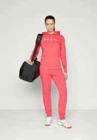 Rózsaszín kapucnis pulóver és illő nadrág fehér logóval, puha anyagból készült. A fekete sporttáska és a fehér sportcipők teszik teljessé az öltözéket.