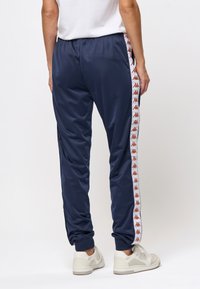 Person som bär marinblå träningbyxor med vita sidostråk som har upprepade röda logotyper, ihop med vita sneakers, stående på vit bakgrund.